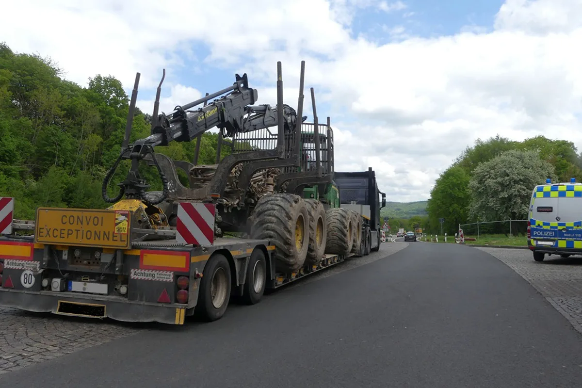 Ein Schwertransporter mit einem großen Fahrzeug beladen steht auf einer Landstraße. Rechts daneben ist ein Polizeiwagen geparkt. Der Himmel ist bewölkt und die Umgebung zeigt grüne Bäume.