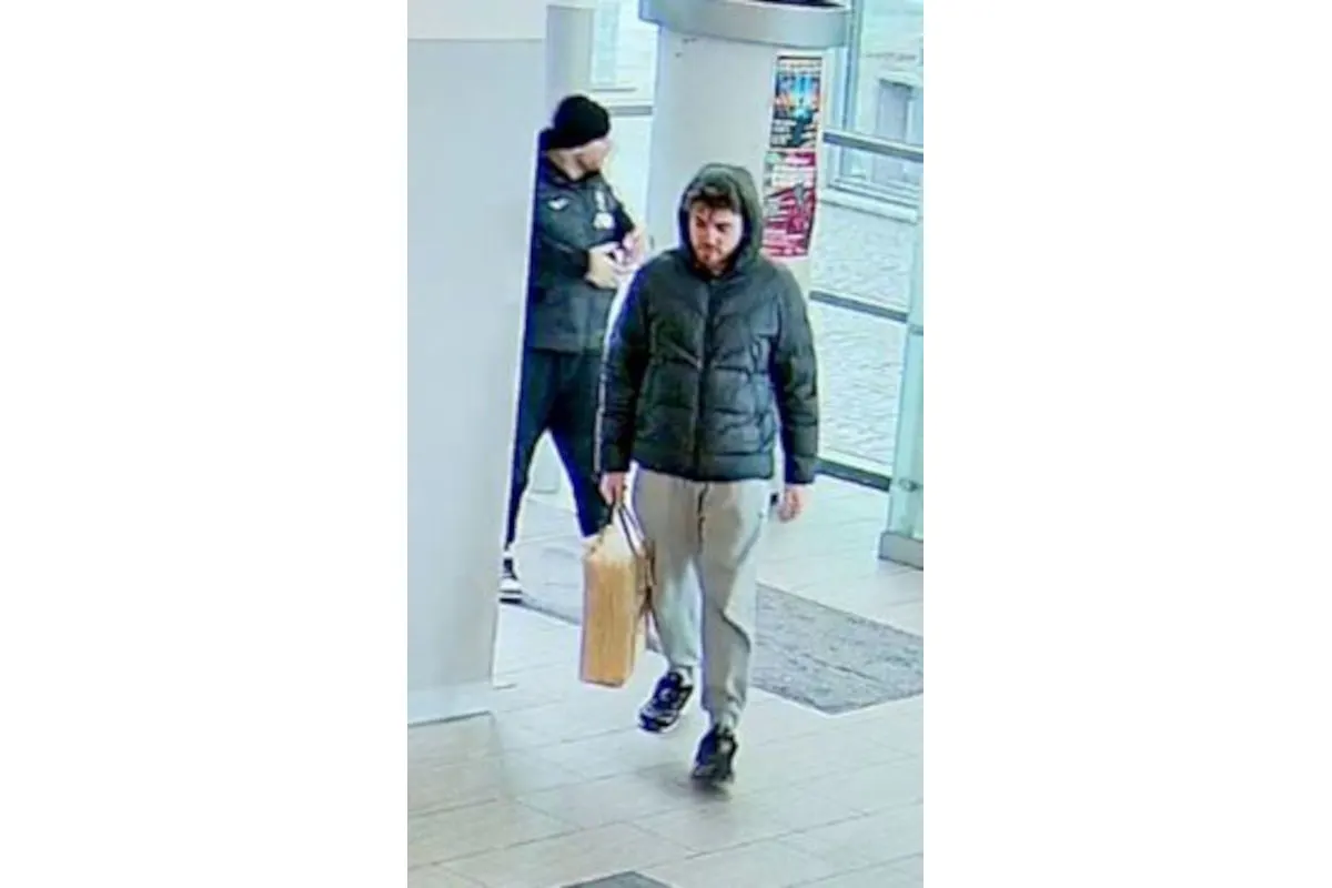 Ein Mann in einer dunklen Jacke, grauer Hose und schwarzen Schuhen trägt eine Tasche in einer Einkaufspassage. Eine weitere Person ist im Hintergrund zu sehen, die etwas in der Hand hält.