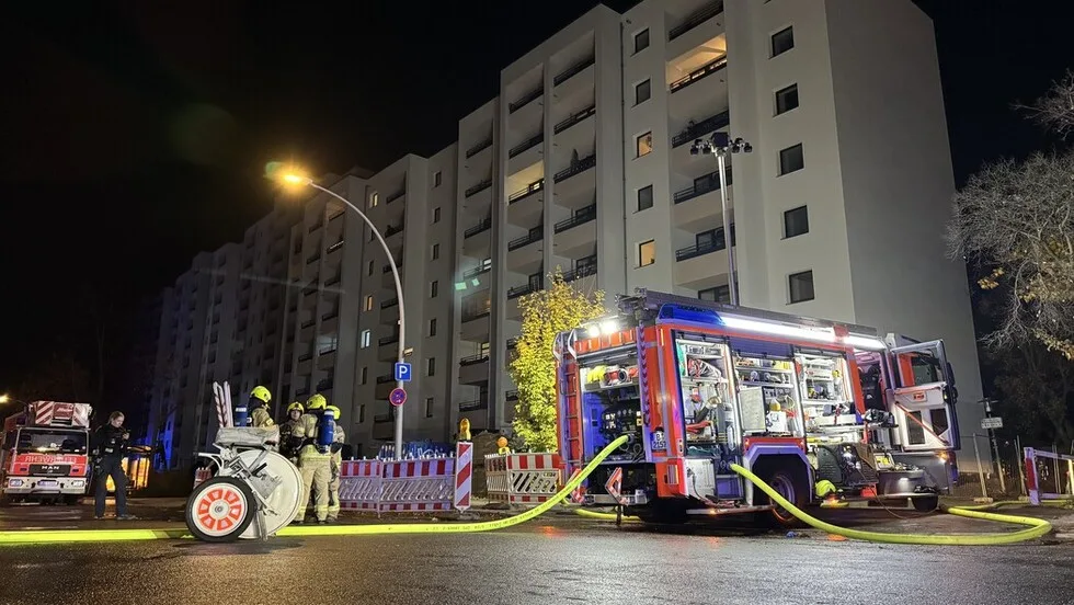 Feuerwehrfahrzeuge und mehrere Feuerwehrleute sind vor einem großen Wohnhaus im Einsatz bei Nacht. Schläuche sind mit dem Fahrzeug verbunden und das Gebäude ist beleuchtet.