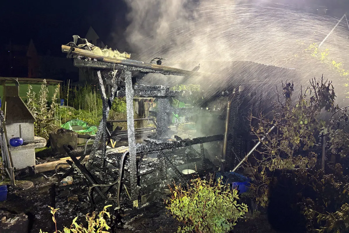 Ein Pavillon in einem Garten ist durch einen Brand völlig zerstört. Überreste sind schwarz verbrannt, und es wird noch gelöscht.