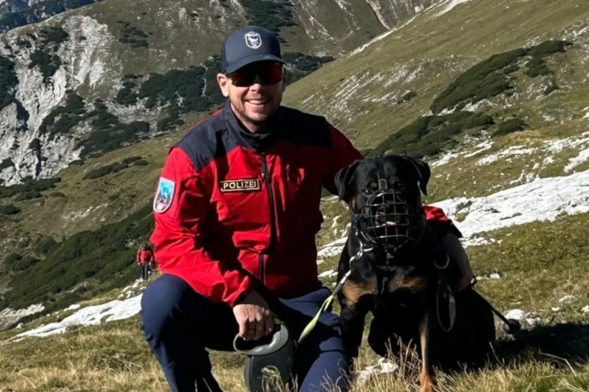 Ein Polizist in roter Uniform ist mit einem Hund, der einen Maulkorb trägt, im alpinen Gelände unterwegs. Im Hintergrund sind Berge und Grünflächen zu sehen.