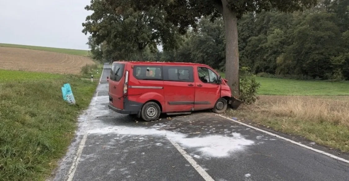 Ein roter Van steht nach einem Unfall frontal gegen einen Baum am Straßenrand. Der Unfallort ist von Streumaterial umgeben.