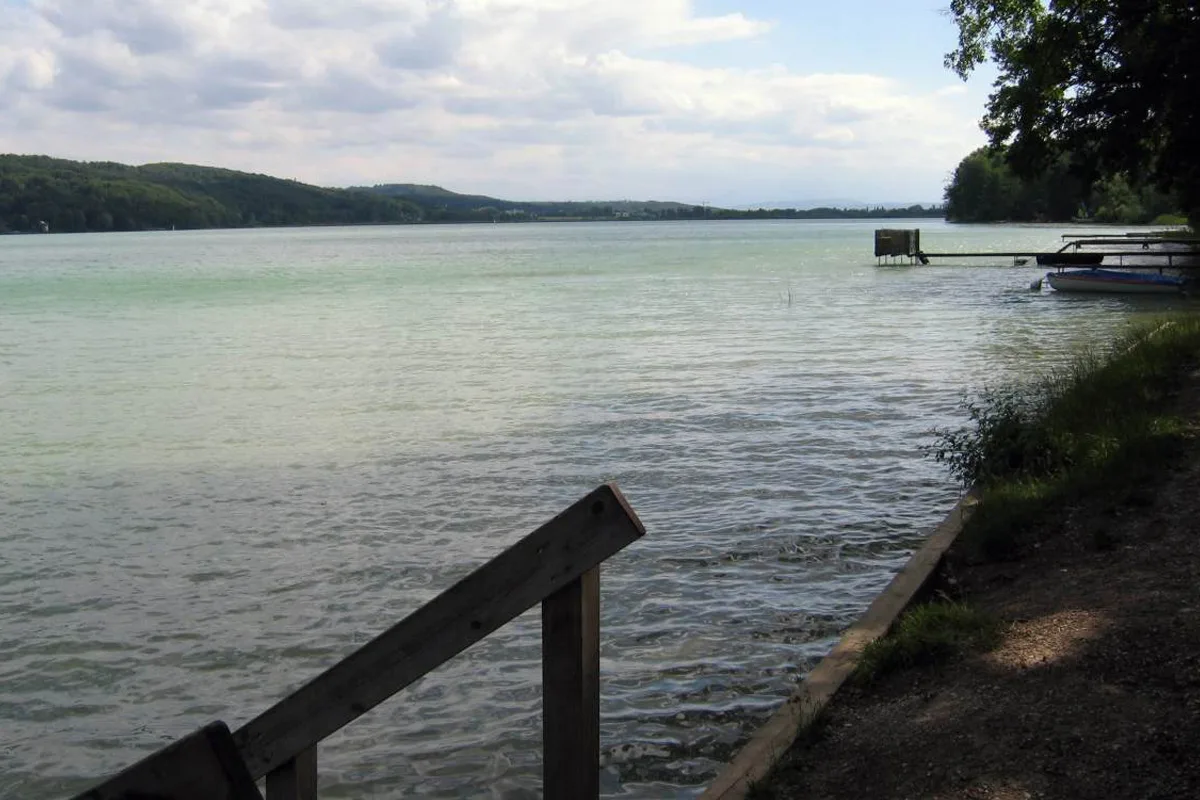 Ein ruhiger See erstreckt sich bis zum Horizont unter einem bewölkten Himmel. Rechts sind Bäume und ein Steg erkennbar.