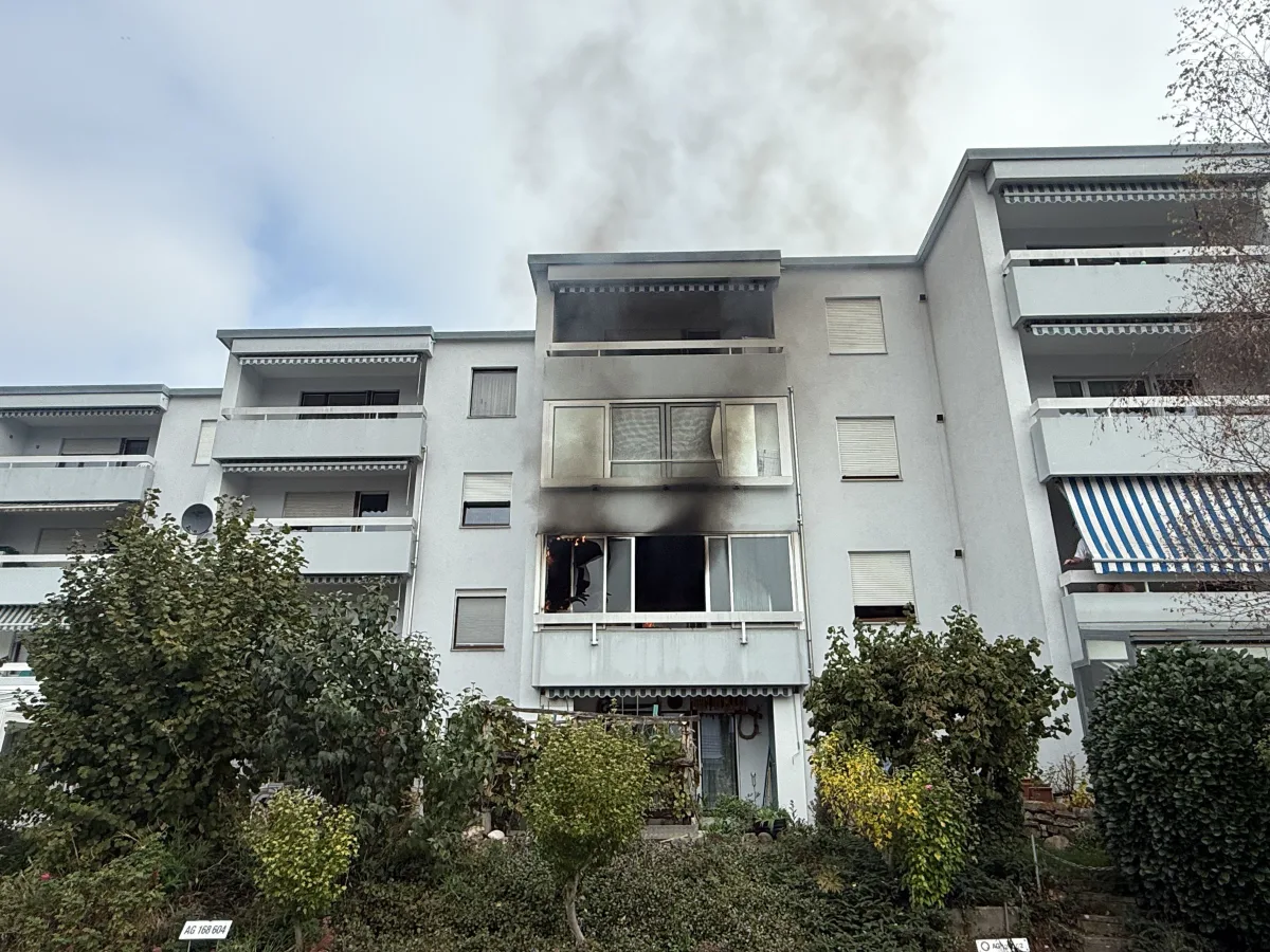 Ein weißes Mehrfamilienhaus, aus dessen Fenster dunkler Rauch aufsteigt. Der Zustand deutet auf ein Feuer hin.