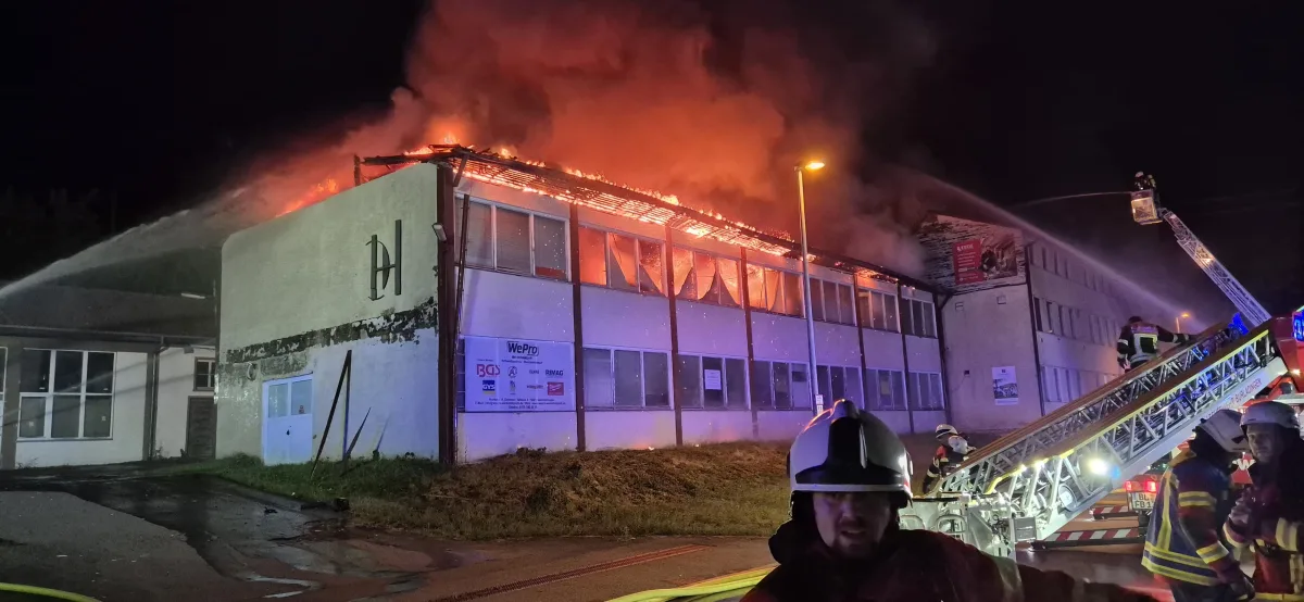 Ein großes Gebäude steht nachts in Flammen, während Feuerwehrleute mit Schläuchen das Feuer bekämpfen. Eine Drehleiter ist im Einsatz.