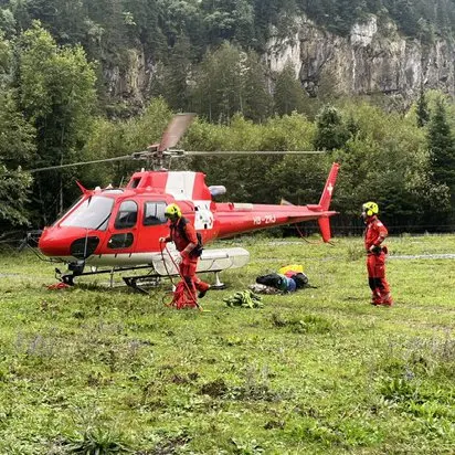 Ein roter Rettungshubschrauber steht auf einer Wiese. Zwei Personen in roten Uniformen sind in der Nähe mit Ausrüstung beschäftigt.