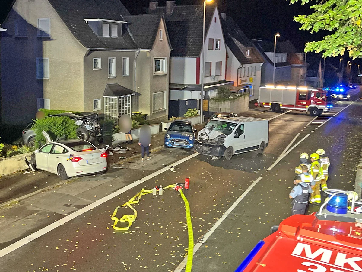 Ein weißes Auto, ein blauer Kleinwagen und ein weißer Van sind nach einem Unfall auf einer Straße stehengeblieben. Feuerwehrleute stehen in der Nähe bereit.