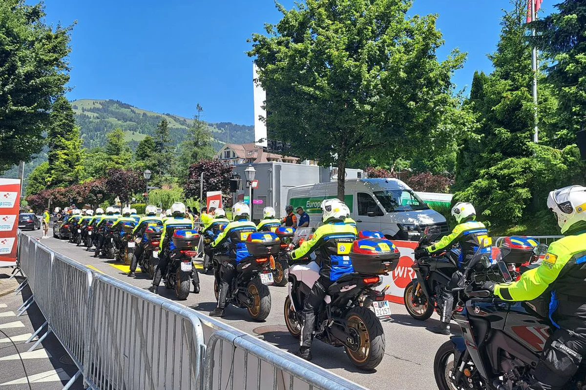 Eine Gruppe von Polizeimotorrädern steht in einer Reihe auf einer abgesperrten Straße. Die Umgebung ist grün und bergig.