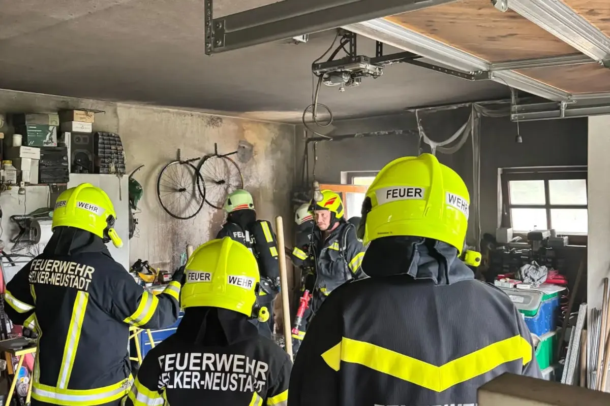 Mehrere Feuerwehrleute in Schutzausrüstung stehen in einer verrauchten Garage mit Fahrrädern und Geräten an den Wänden.