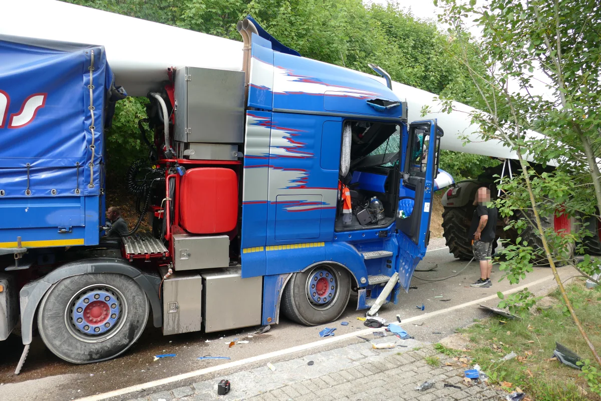 Ein blauer Lastwagen zeigt deutliche Unfallschäden an der Seite. Im Hintergrund ist ein großer Traktor zu sehen, der am Unfall beteiligt zu sein scheint.