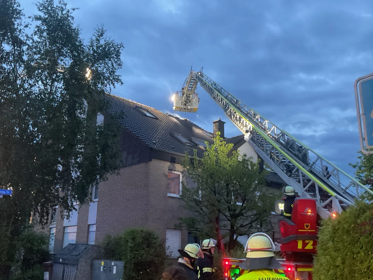 Mehrere Feuerwehrleute in Schutzkleidung arbeiten mit einer Drehleiter an einem Hausdach. Es ist dämmerig.