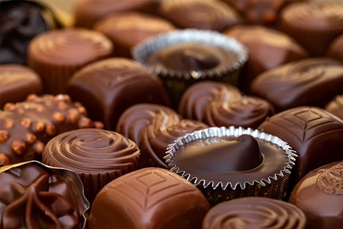 Das Bild zeigt eine Nahaufnahme einer Vielzahl von Pralinen, die in einem Schaufenster präsentiert werden. Die Pralinen variieren in Form und Größe, mit unterschiedlichen Mustern und Farben aus dunkler und Milchschokolade.