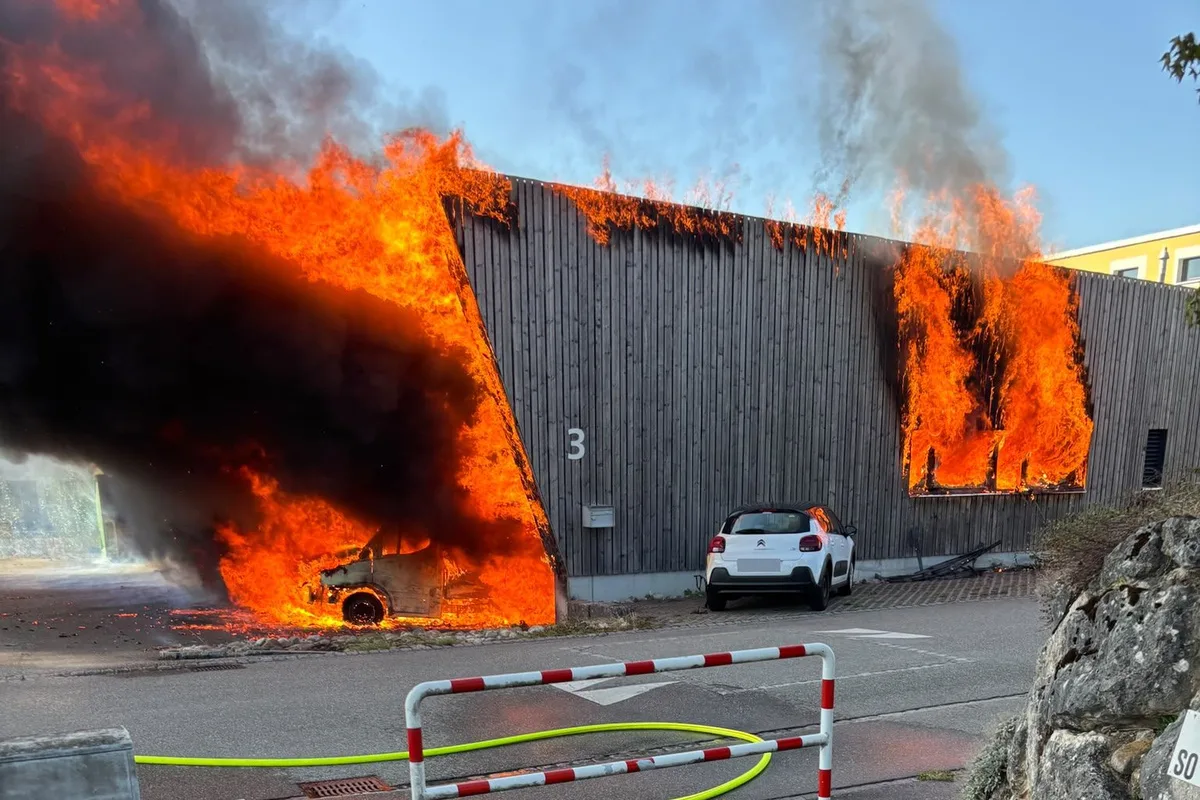 Ein Fahrzeug brennt neben einem Gebäude, das ebenfalls in Flammen steht. Schwarzer Rauch steigt in den Himmel.