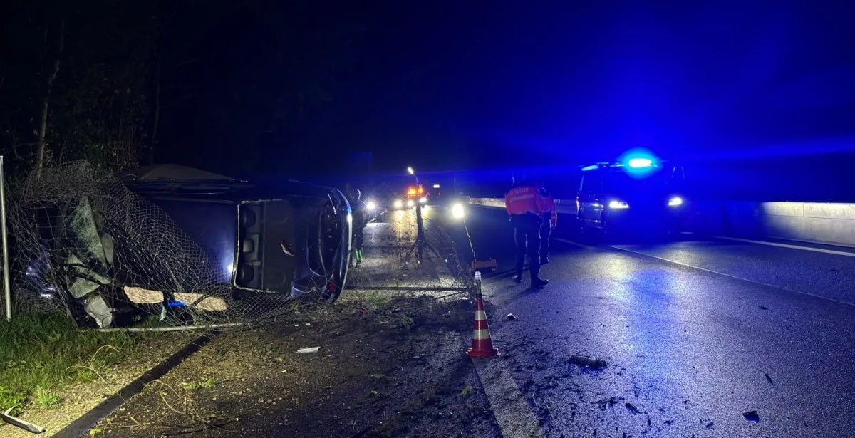 Ein Auto liegt bei Nacht auf der Seite neben einem beschädigten Zaun einer Autobahn. Polizeifahrzeuge mit Blaulicht sind im Hintergrund sichtbar.