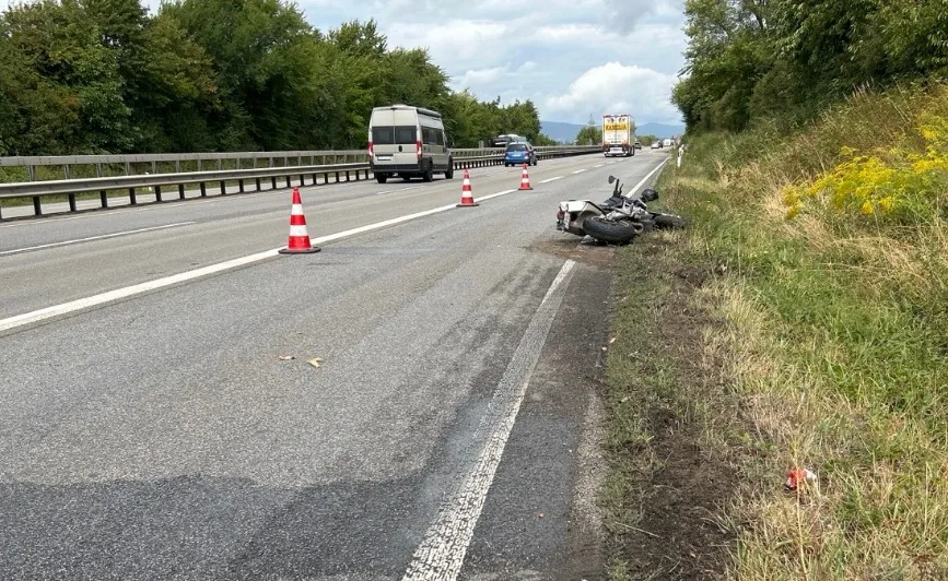 Ein verunfalltes Motorrad liegt neben einer Landstraße. Verkehrshütchen sind aufgestellt, und ein Van fährt vorbei.