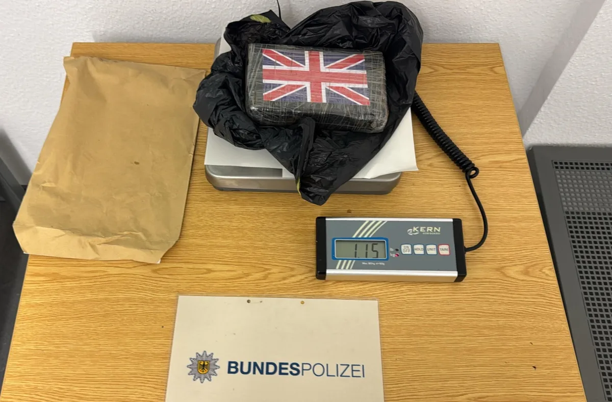 Ein rechteckiges Paket mit der britischen Flagge liegt auf einer Waage. Daneben ein Schild mit 'BUNDESPOLIZEI'.