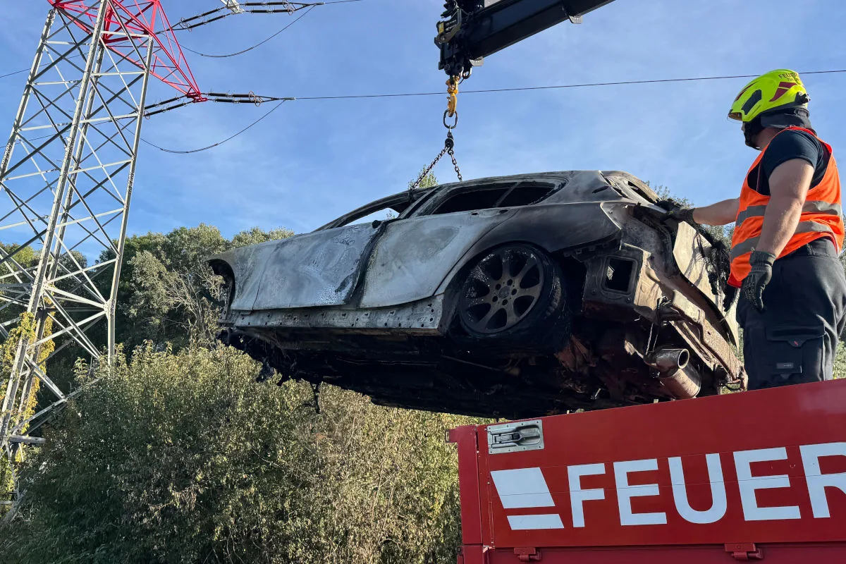 Ein Feuerwehrmann hebt ein ausgebranntes Auto mit einem Kran an. Im Hintergrund steht ein großer Strommast. Der Himmel ist klar.
