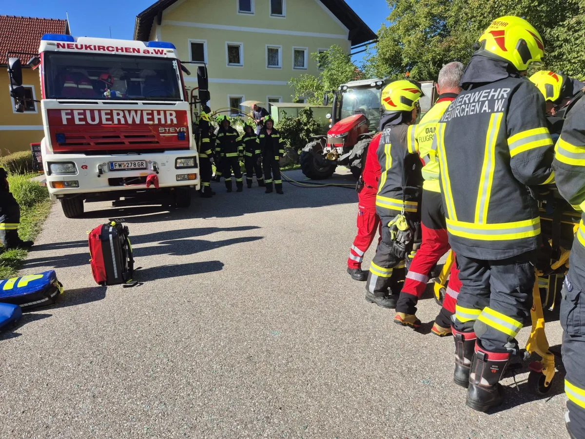 Mehrere Feuerwehrleute in Uniform stehen neben einem großen roten Feuerwehrfahrzeug der Feuerwehr Neukirchen a.W. Ein roter Traktor ist ebenfalls vor Ort neben einem gelben Haus zu sehen.
