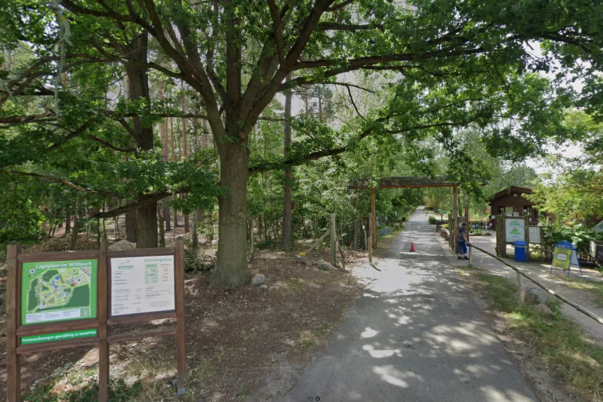 Der Eingang eines Wildparks zeigt mehrere Bäume, Schilder mit Informationen und einen Weg in den Park.