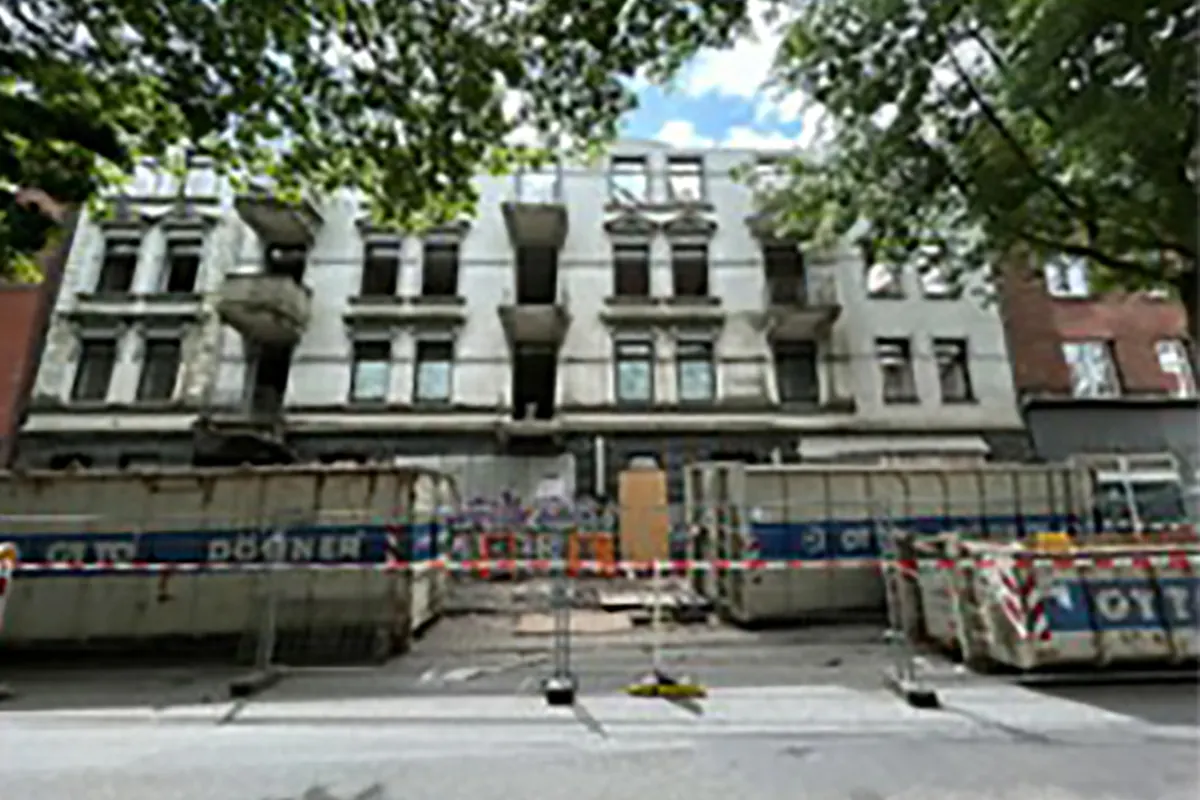 Ein mehrstöckiges Gebäude in einer urbanen Umgebung, dessen Fassade sichtbare Schäden zeigt. Absperrungen und Baucontainer befinden sich davor.