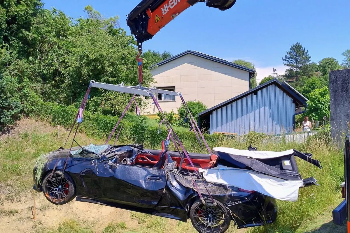 Ein stark beschädigtes, schwarzes Auto mit rotem Innenraum wird von einem Kran in einem grünen Wohngebiet geborgen. Im Hintergrund sind Bäume und Wohngebäude zu sehen.