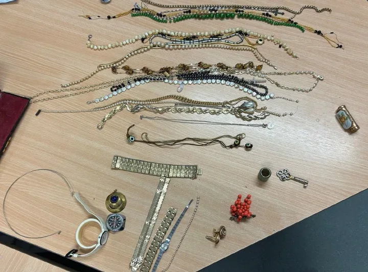 Diverse Schmuckstücke, darunter Ketten, Armbänder und Uhren, sind auf einem Holztisch verteilt zu sehen.