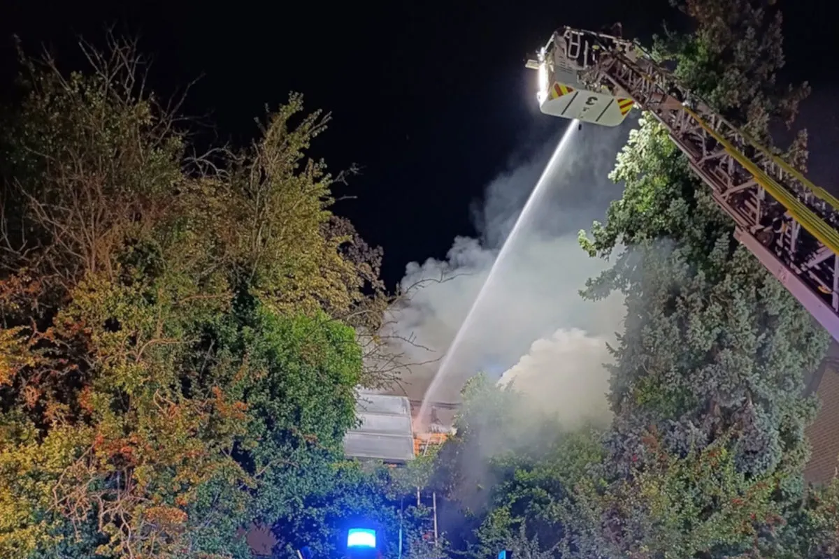Eine Feuerwehrleiter sprüht Wasser auf ein brennendes Gebäude, das sich hinter Bäumen versteckt. Rauch steigt auf.