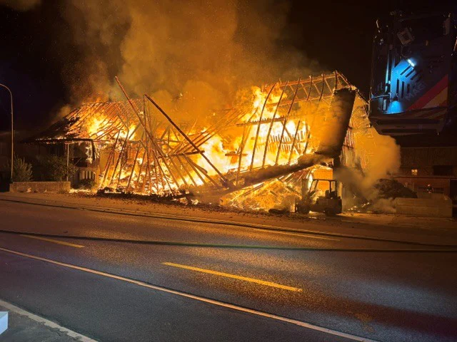 Ein Gebäude steht in Flammen, das Dach ist bereits eingestürzt. Die Straße verläuft parallel zu dem brennenden Gebäude, während dichter Rauch aufsteigt.