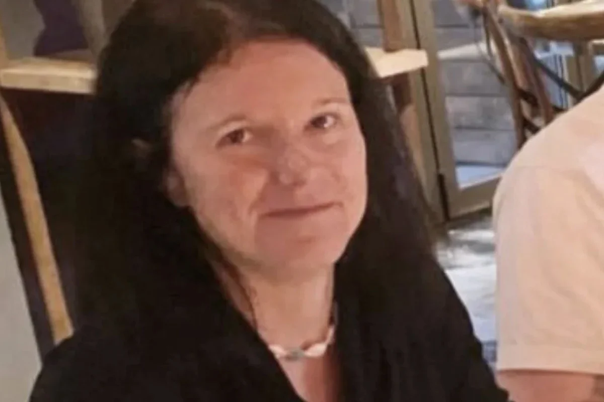 Eine Frau mit dunkelbraunem Haar sitzt drinnen und schaut zur Kamera. Sie trägt eine schwarze Oberbekleidung.