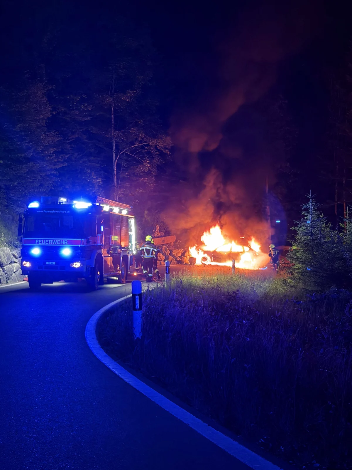 Ein Feuerwehrfahrzeug mit eingeschaltetem Blaulicht steht auf einer Landstraße. Davor brennt ein Auto, umgeben von Feuerwehrleuten, in einer nächtlichen Waldszenerie.