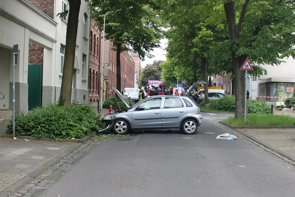 Ein silberfarbener Wagen mit erheblichen Frontschäden steht quer auf einer Straßenkreuzung in einer Wohngegend. Im Hintergrund sind Einsatzkräfte und Fahrzeuge der Rettungsdienste zu sehen.