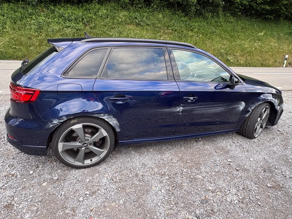 Ein blauer Audi ist am Straßenrand geparkt, mit deutlichen Schrammen an der rechten Fahrzeugseite, einer grünen Böschung im Hintergrund.