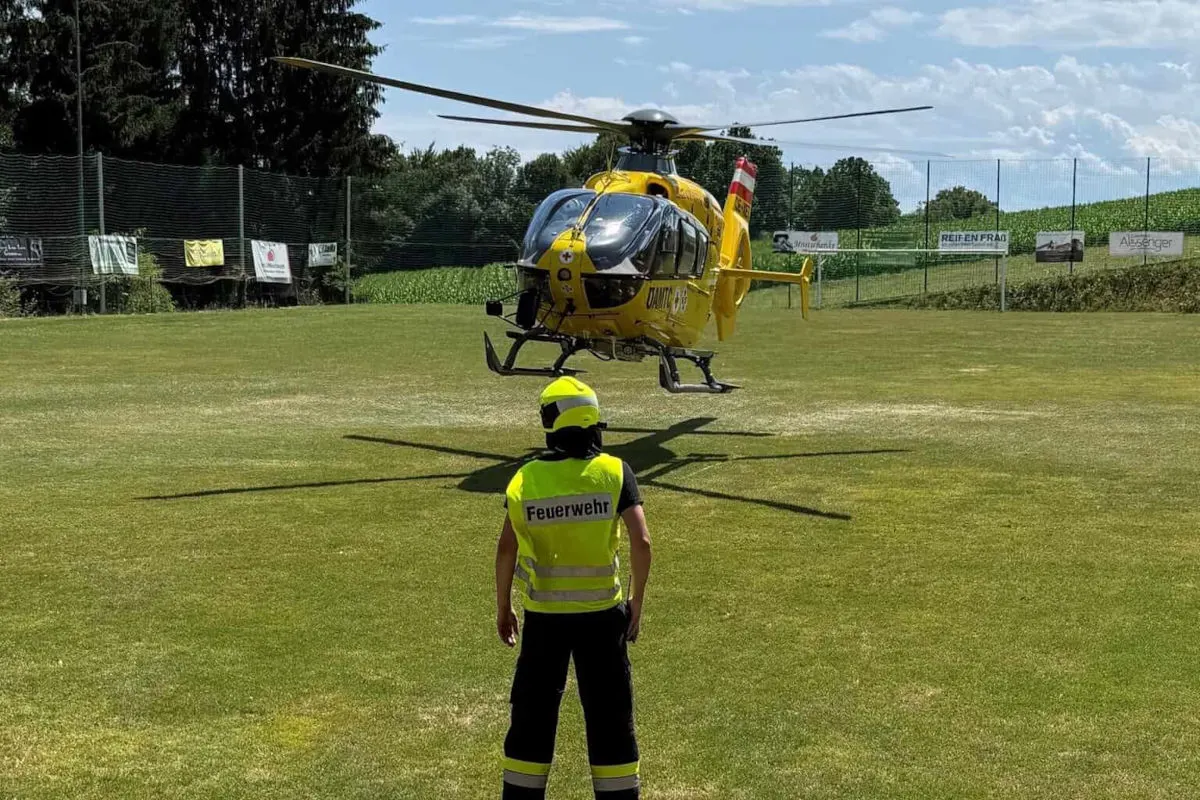 Ein Feuerwehrmann beobachtet einen gelben Rettungshubschrauber, der auf einem grünen Feld landet. Im Hintergrund sind Bäume und ein Zaun sichtbar.