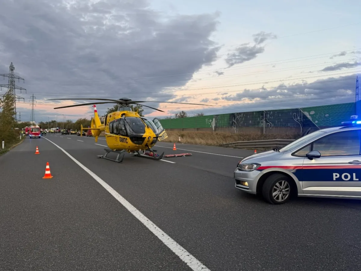 Ein gelber Rettungshubschrauber ist auf einer Autobahnstraße gelandet, umgeben von Absperrungen und einem Polizeiauto. Die Szene spielt sich während der Dämmerung ab.