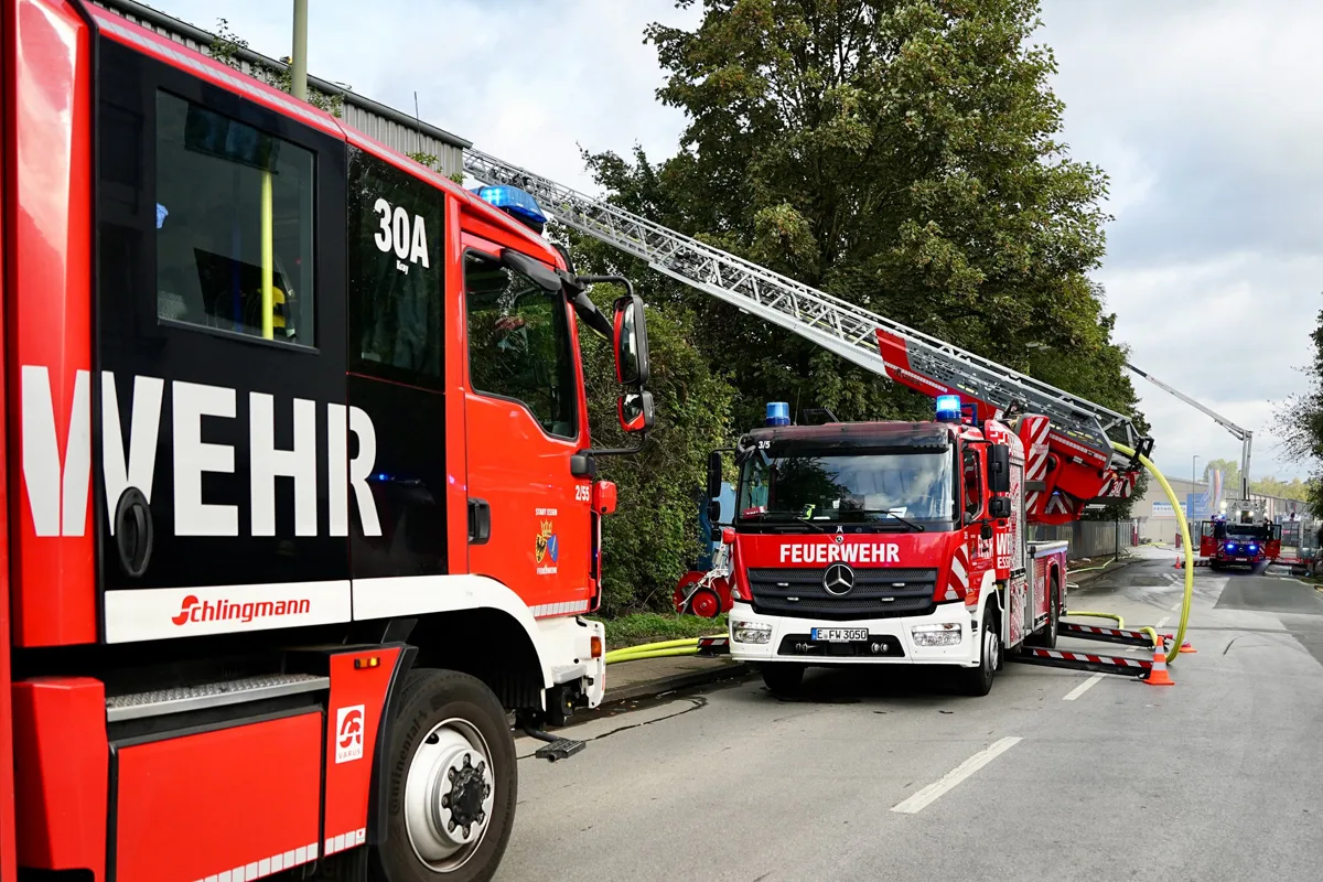 Drei Feuerwehrfahrzeuge stehen auf einer städtischen Straße, eines davon mit ausgefahrener Drehleiter. Es sind Bäume und ein Gebäude im Hintergrund zu sehen.