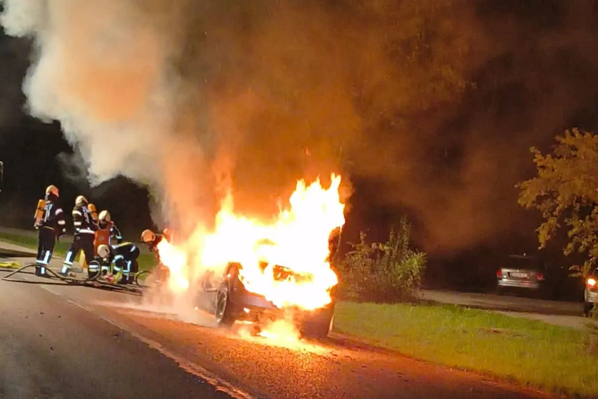 Bei Nacht bekämpfen Feuerwehrleute einen Fahrzeugbrand auf einer Straße. Flammen schlagen aus der Motorhaube des Fahrzeugs.