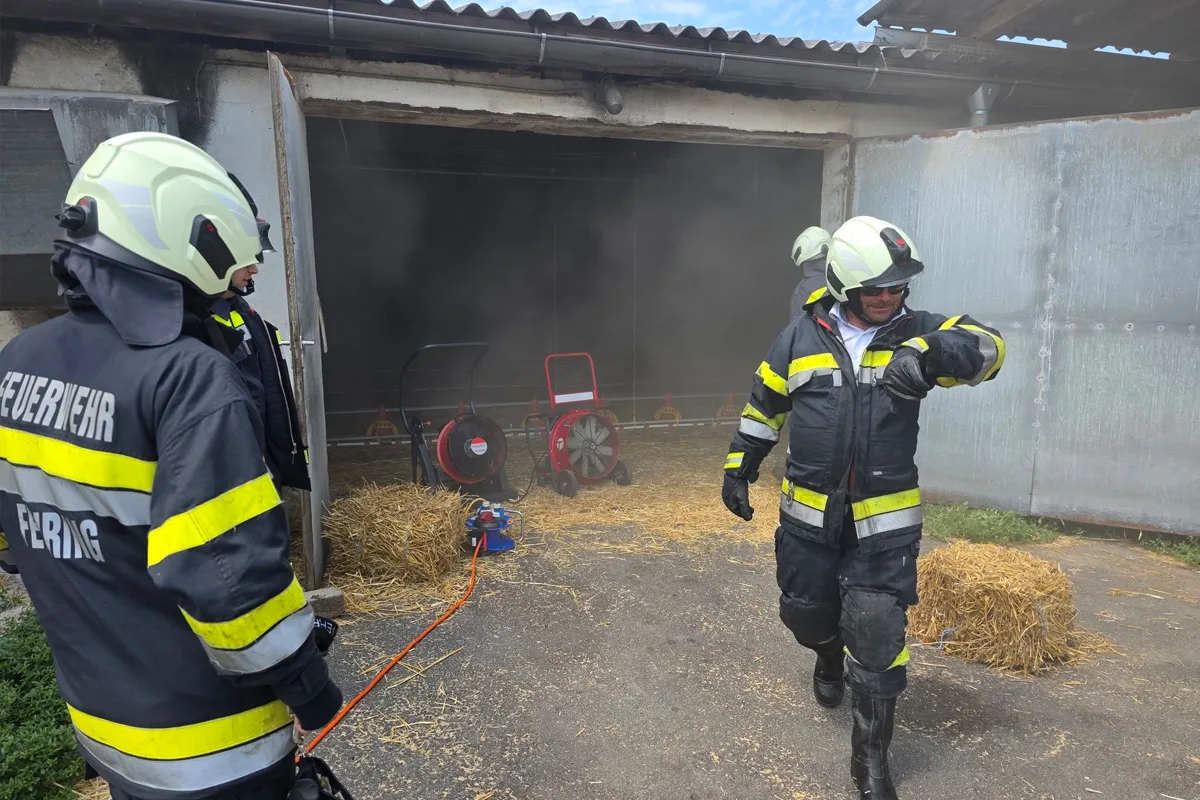 Mehrere Feuerwehrleute stehen vor einem verrauchten Gebäude. Zwei Lüfter sind zur Belüftung aufgestellt.