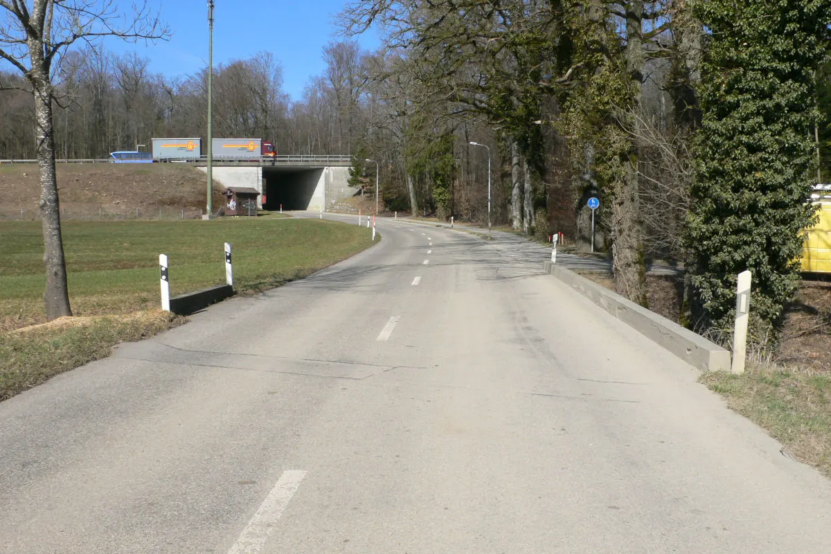 Eine gerade Landstraße führt durch eine ländliche Gegend in Richtung einer Unterführung. Kein Verkehr ist sichtbar, und der Himmel ist klar.