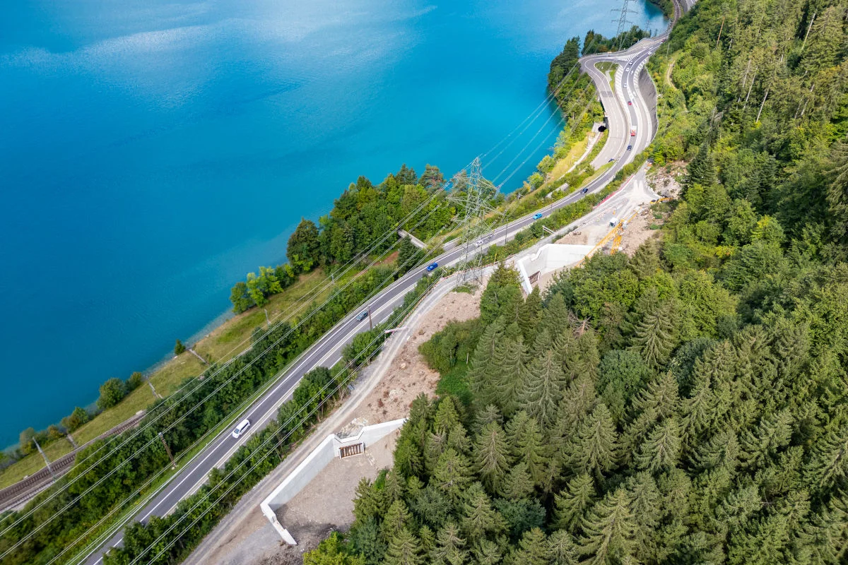 Eine kurvige Straße verläuft entlang eines blauen Sees, umgeben von dichtbewaldeten Hängen. Stromleitungen sind sichtbar.