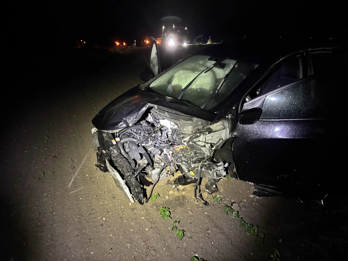 Ein stark beschädigtes Auto mit erheblichen Frontschäden steht nachts auf einer unbefestigten Straße. Im Hintergrund sind Lichter von Einsatzfahrzeugen zu sehen.