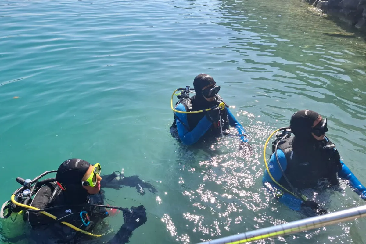 Drei Taucher, die komplette Tauchausrüstung tragen, stehen in einem Gewässer im Freien. Die Sonne scheint und das Wasser wirkt klar.