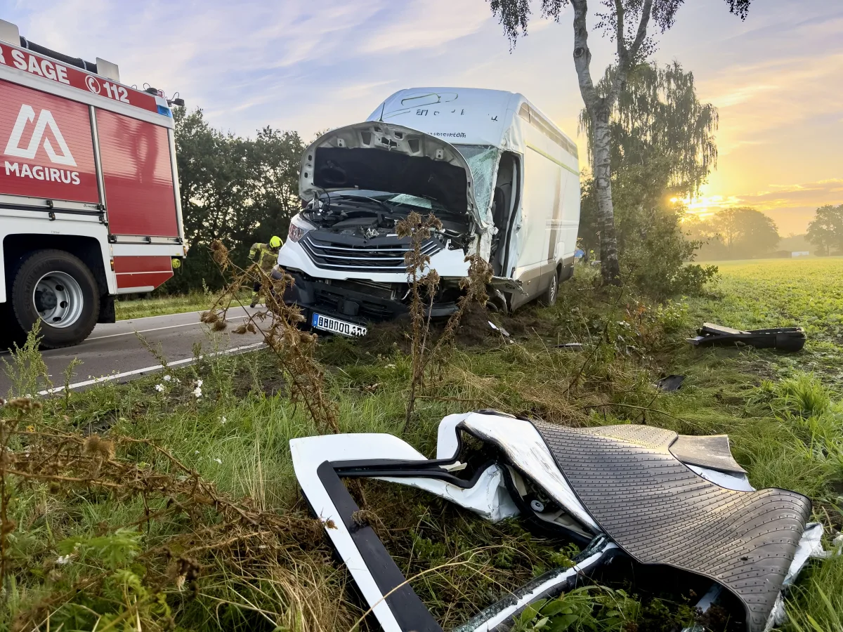 Ein weißer Lieferwagen ist stark beschädigt und steht neben einem Baum. Ein Feuerwehrauto ist auf der Straße sichtbar. Die Szene ist bei Sonnenaufgang.