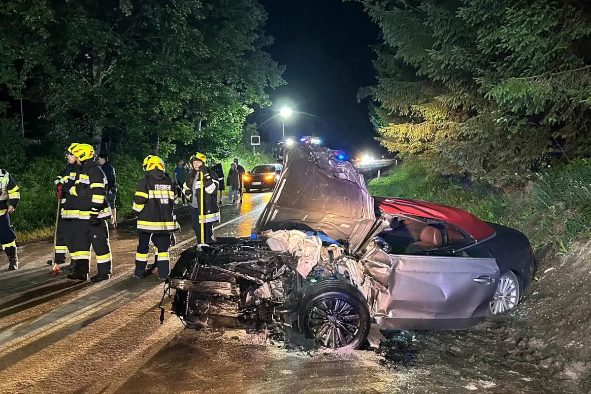 Ein stark verunfalltes Auto steht auf einer Landstraße bei Nacht. Mehrere Feuerwehrleute in Arbeit sind zu sehen, einige Personen stehen im Hintergrund.