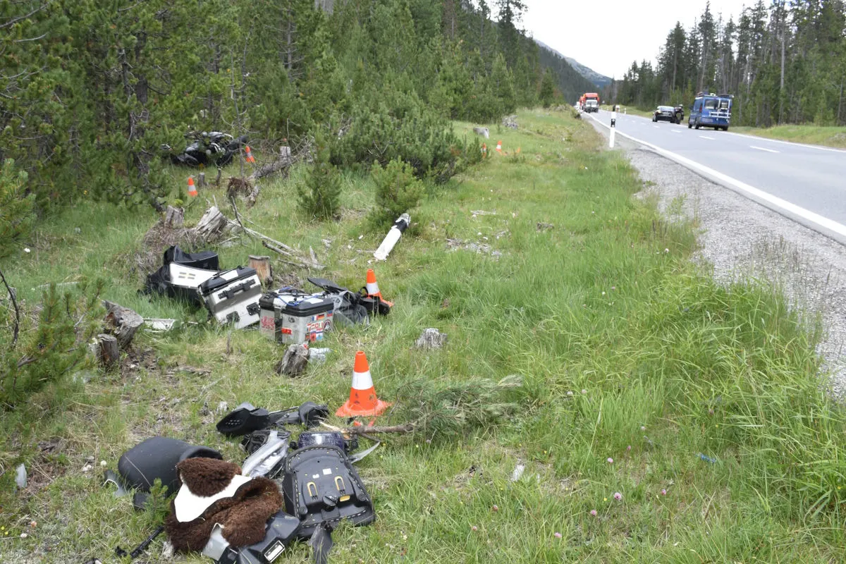 Auf einem Grünstreifen neben einer Landstraße liegen verstreute Motorradteile und Gepäckstücke. Zwei Fahrzeuge sind auf der Straße sichtbar.