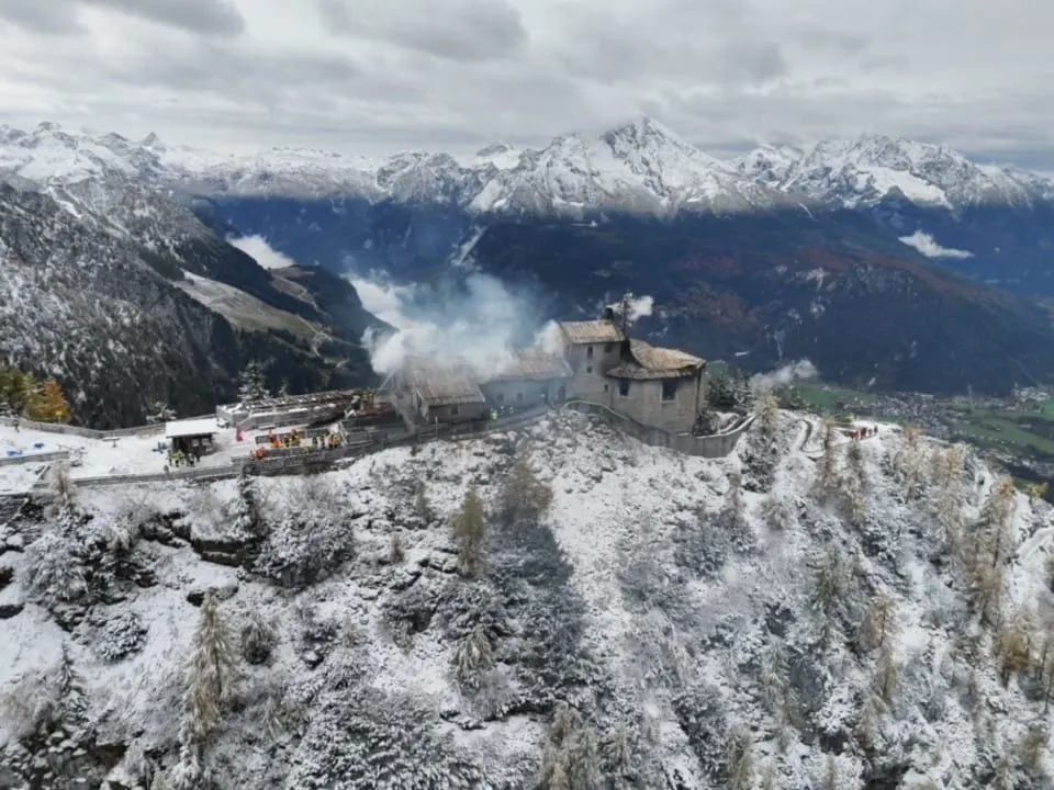 Ein historisches Gebäude, das Kehlsteinhaus, steht auf einem verschneiten Berg, während Rauch aufsteigt. Im Hintergrund sind verschneite Berge zu sehen.