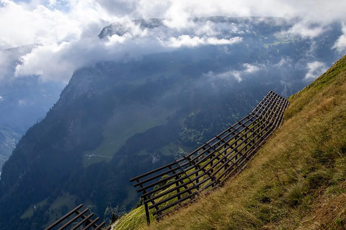 Ein steiler Hang in den Alpen mit einer Lawinenverbauung im Vordergrund, umgeben von Wolken und einem Bergpanorama.