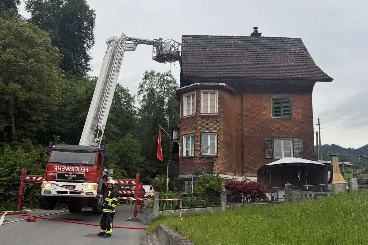 Ein Feuerwehrfahrzeug mit Drehleiter steht auf einer Straße neben einem roten Wohnhaus. Ein Feuerwehrmann in Uniform ist ebenfalls zu sehen.