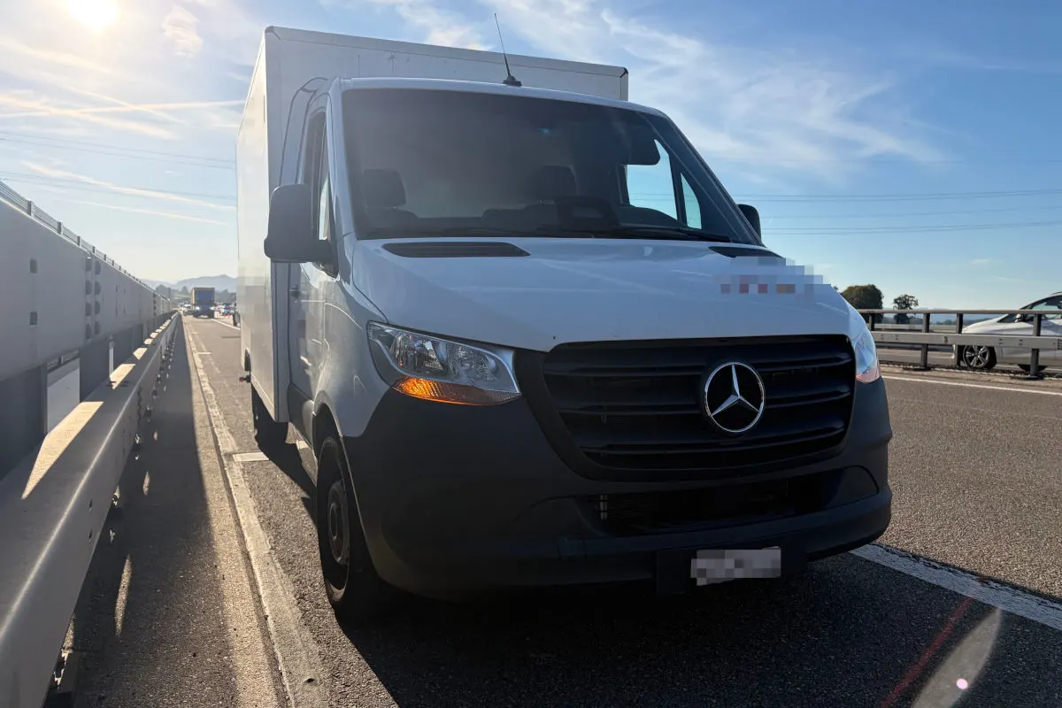 Ein weißer Mercedes-Transporter steht auf einem Seitenstreifen einer Autobahn. Im Hintergrund sind weitere Fahrzeuge und eine Leitplanke zu sehen. Der Himmel ist überwiegend klar.
