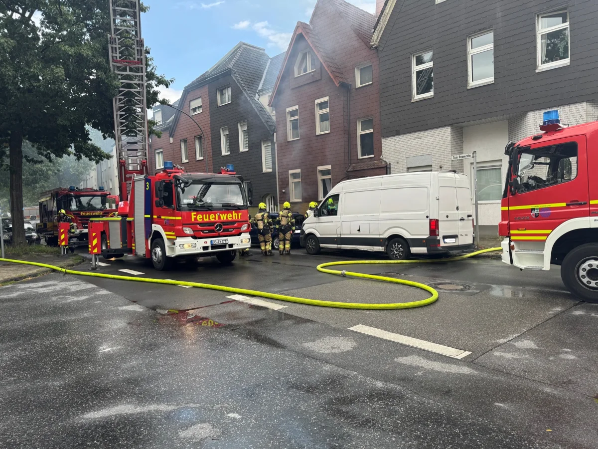 Mehrere Feuerwehrfahrzeuge stehen vor einem Wohnhaus, während Feuerwehrleute mit einem Schlauch die Situation kontrollieren.
