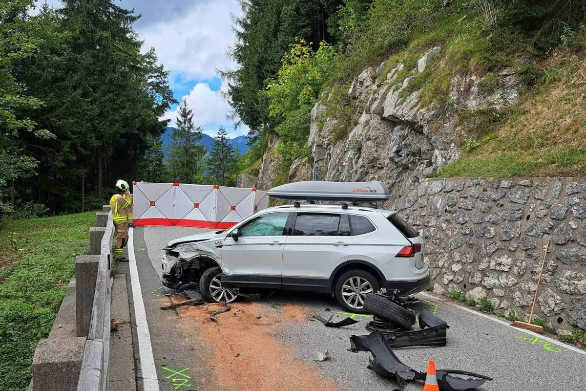 Ein SUV ist nach einem Unfall stark beschädigt, umgeben von Trümmern auf einer schmalen Bergstraße. Ein Einsatzkraft in Schutzkleidung steht daneben.